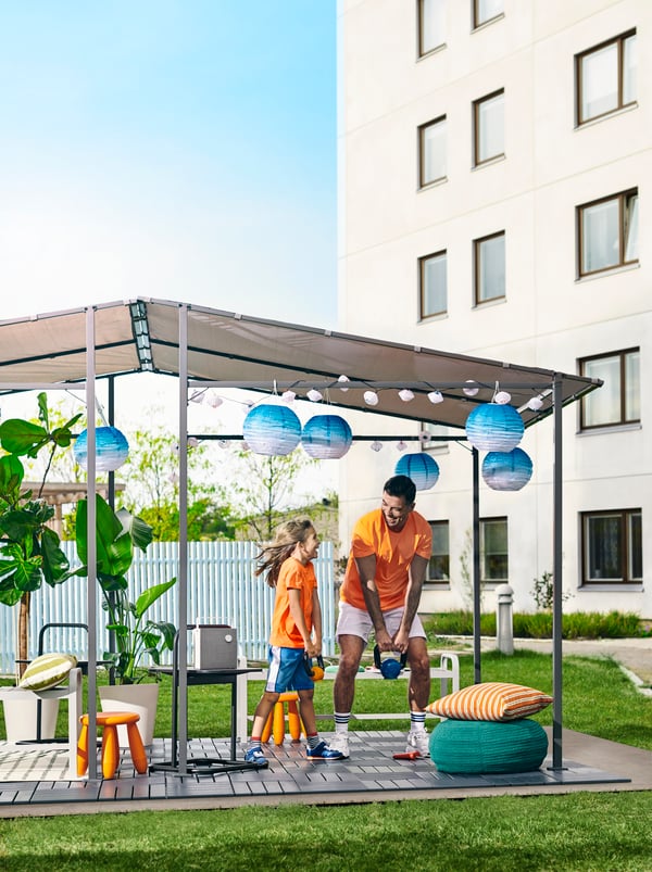 A man and a child listen to music from an ENEBY Bluetooth speaker and lift weights inside a GUNNÖN gazebo in a park.
