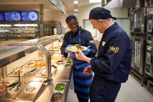 Deux employés en uniforme bleu foncé avec « Hej ! » sur les manches lors d’un buffet en libre-service dans un restaurant d’entreprise ; L’un montre une assiette de nourriture à l’autre, avec une vue sur les plats chauds, les fours et les assiettes de menu en arrière-plan.