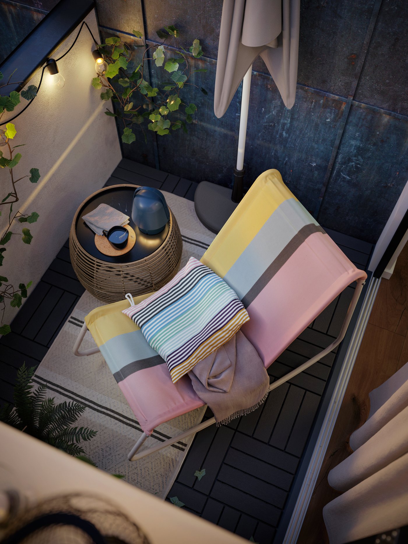 Une chaise berçante TUMHOLMEN blanc et multicolore à côté d’une table basse d’extérieur sur un tapis, plus une guirlande lumineuse et une plante.
