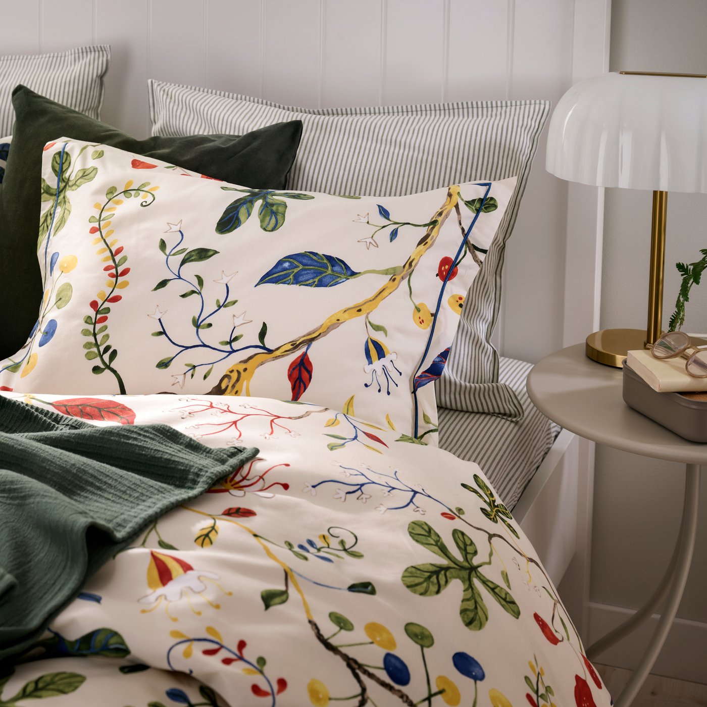 Close up of a bed featuring KRONBUSKE duvet and pillow cover and a green blanket, and a small bedside table.