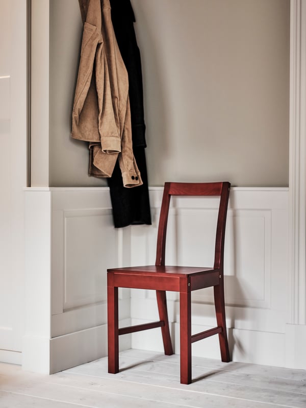 A corner of a room with beige and black items of clothing hanging from a wall next to a red stained PINNTORP chair.