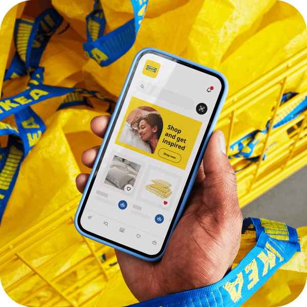 A hand holds a smartphone displaying the IKEA app, surrounded by yellow IKEA bags with blue handles in a shopping cart