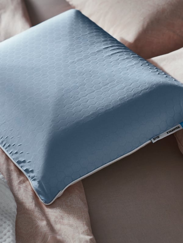 A KVARNVEN ergonomic pillow, with a cooling blue side with a honeycomb pattern, lies on top of a bed.