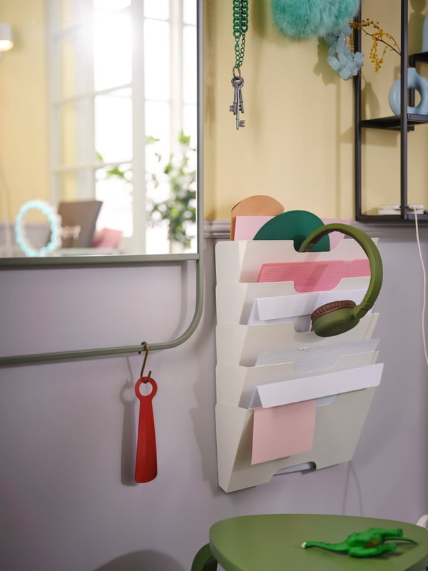 Väggmonterad dokumenthållare med papper och mappar. Gröna hörlurar hänger på toppen, rött skohorn på sidan. Solljus från fönster i bakgrunden.