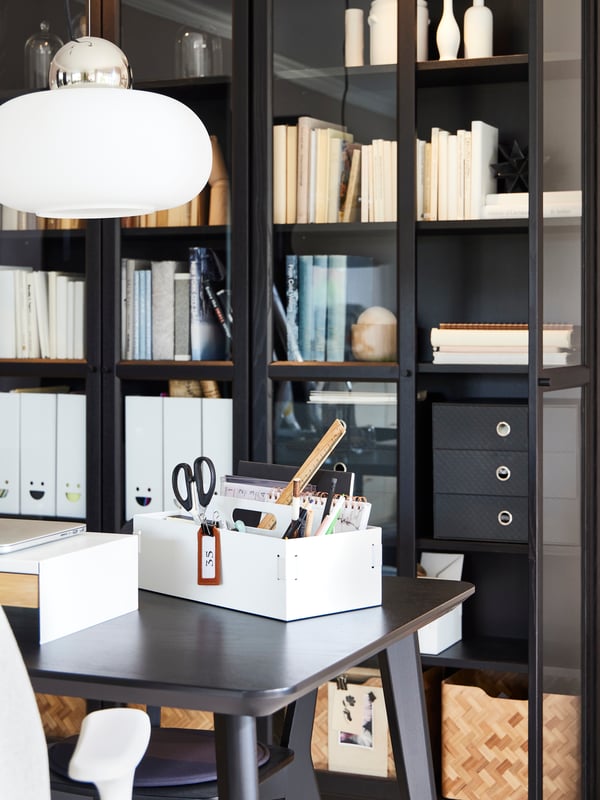 One end of a dining table in front of glass-door bookcases, with a KVISSLE desk organiser on top holding work accessories.