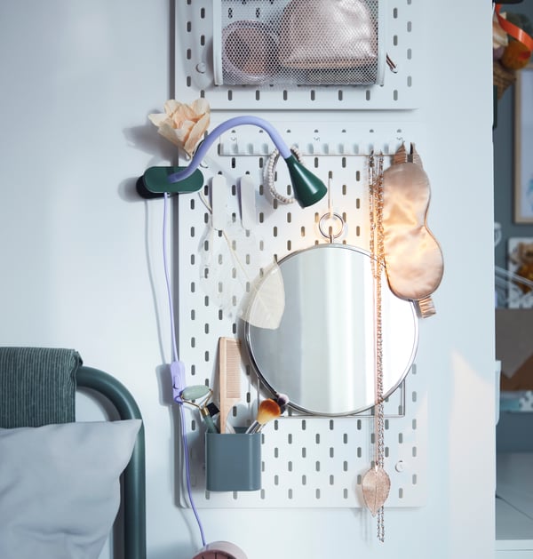 Miroir rond et récipients suspendus à un panneau perforé SKÅDIS blanc, où sont rangés du matériel de maquillage, des bijoux et des accessoires.