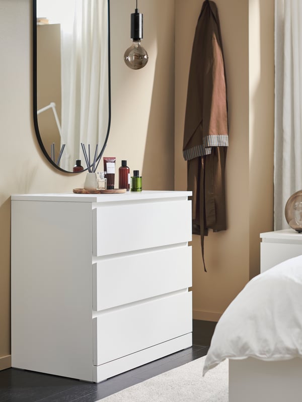 A white MALM chest with 3 drawers against a beige wall with bottles and other objects on top and a large mirror behind it.