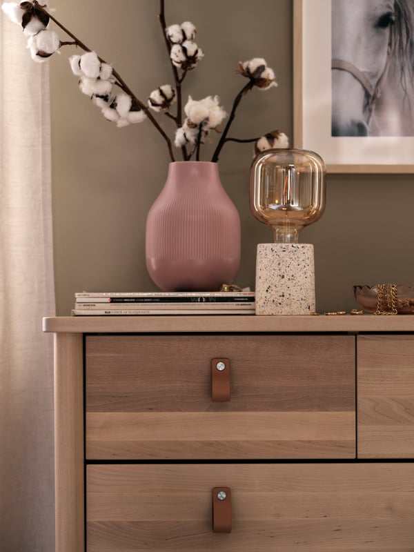 A pink GRADVIS vase with blooming twigs and a LERSKIFFER lamp with a LUNNOM LED bulb stand on top of a chest of drawers.