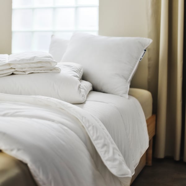 Sur un lit, deux couettes SKOGSFRÄKEN pliées sur une autre dépliée, toutes sans housse, à côté de deux oreillers.