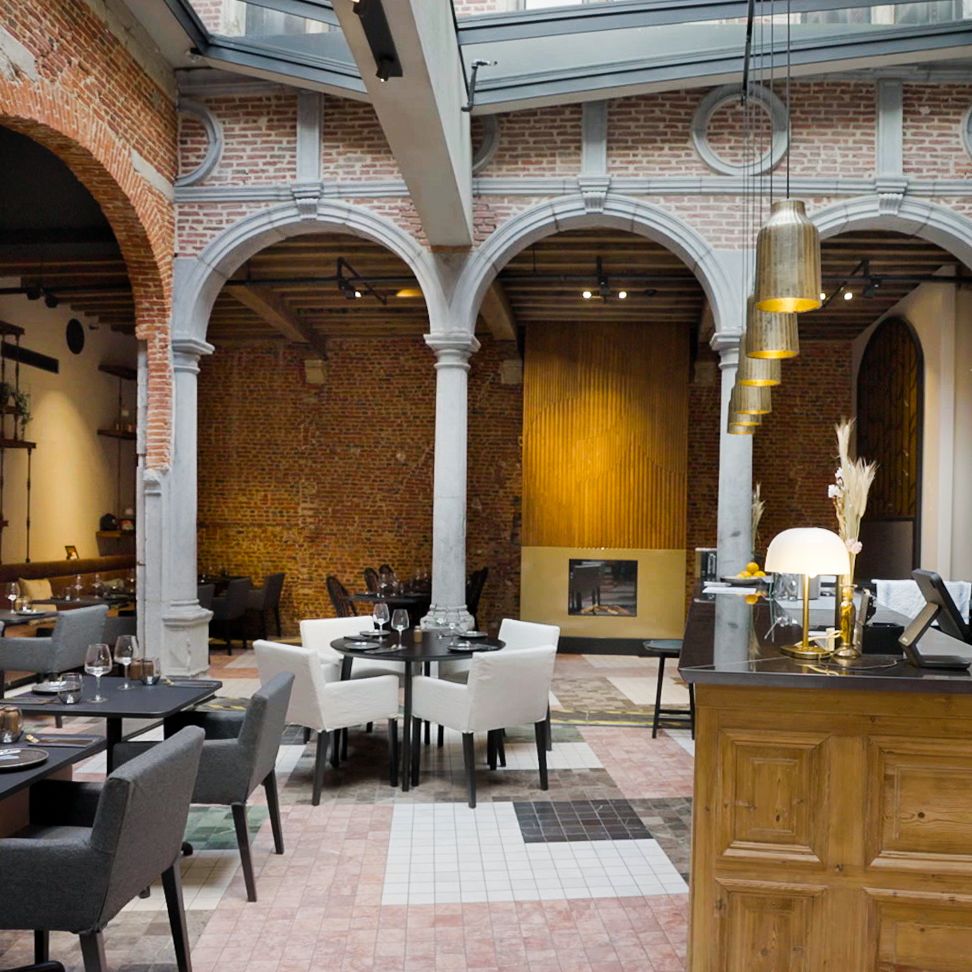 Restaurant met hoge glazen dak, bakstenen muren en bogen. Zwarte en witte tafels met stoelen, houten balie en gouden hanglampen in stijlvol decor.