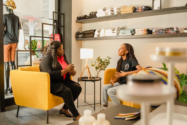 Two people sit opposite each other in a store environment, surrounded by clothes on shelves and clothes racks, with a small table and lamp between them.