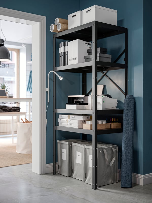 A black BROR shelving unit is filled with office supplies. A NÄVLINGE LED wall spotlight illuminates a printer on one shelf.