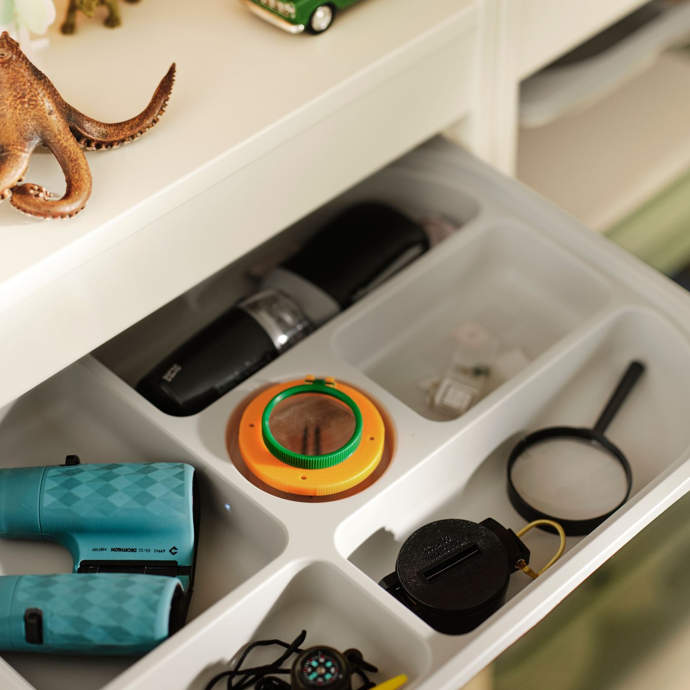 Un cassetto bianco aperto contiene tanti piccoli oggetti suddivisi nei vari scomparti. In alto ci sono degli animali giocattolo.