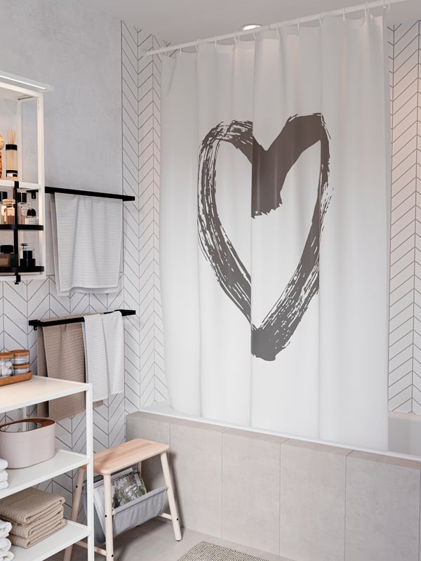 A white shower curtain with a big grey heart illustration in the middle. Black towel rails with white and beige towels.