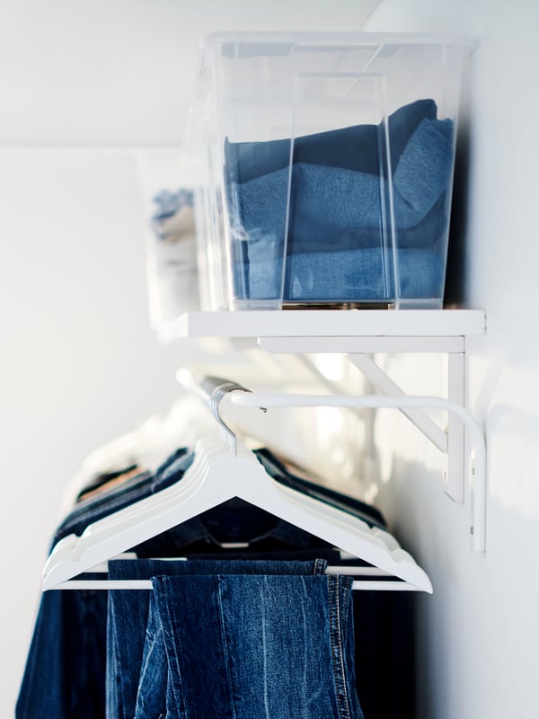 Un colgador de ropa MULIG con prendas de colores azules colgadas de ganchos blancos. Sobre el colgador hay una repisa blanca con cajas transparentes SAMLA donde se ve ropa guardada.