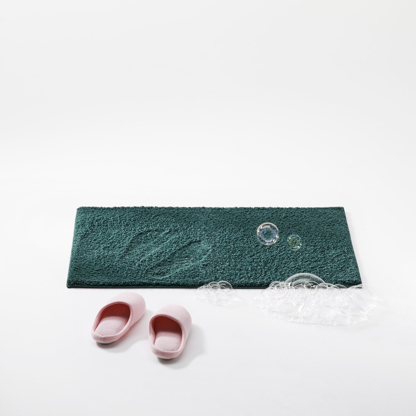 Pink slippers and soap bubbles resting beside a BACKMÅRA bath mat in a clean white studio setting.