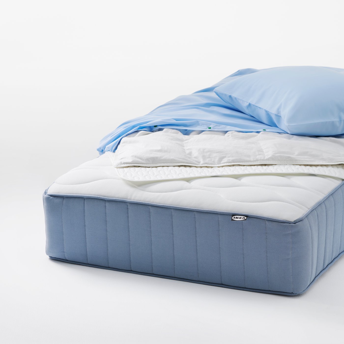 An uncovered VÅGSTRANDA pocket sprung mattress with light bedding placed on top in a clean studio setting.
