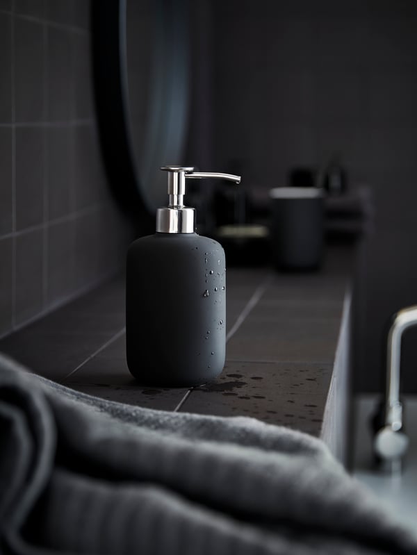 A dark grey EKOLN soap dispenser is standing in a grey bathroom, next to a mirror and with a grey towel in the foreground.