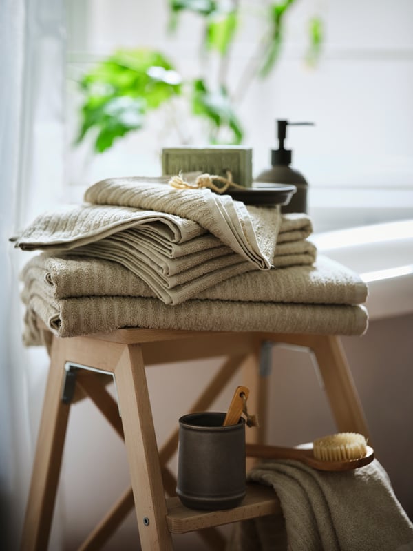 A pile of VÅGSJÖN cotton towels in beige colour on a wooden stool in front of a window. Soap and a body brush next to it.  