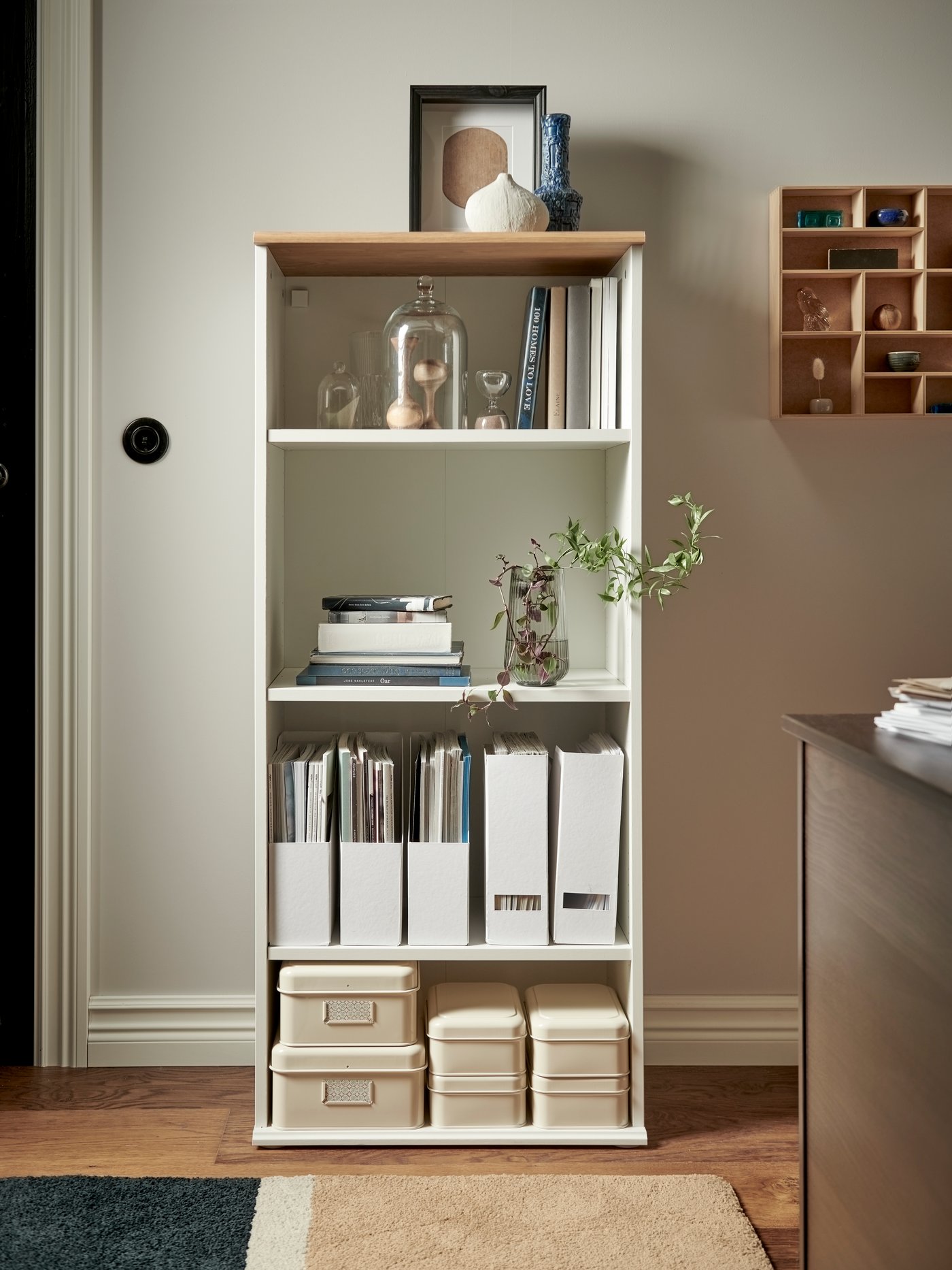 A white SKRUVBY bookcase with decorative items, storage boxes with lids and magazine files stands against a white wall.