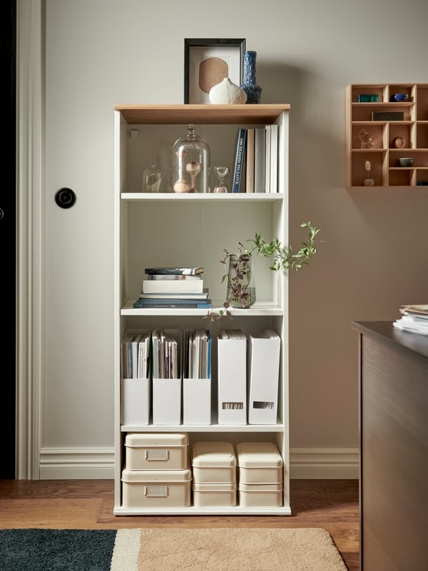 Una SKRUVBY librería blanca con decorativo artículos, cajas de almacenaje con tapa y revisteros contra una pared blanca.