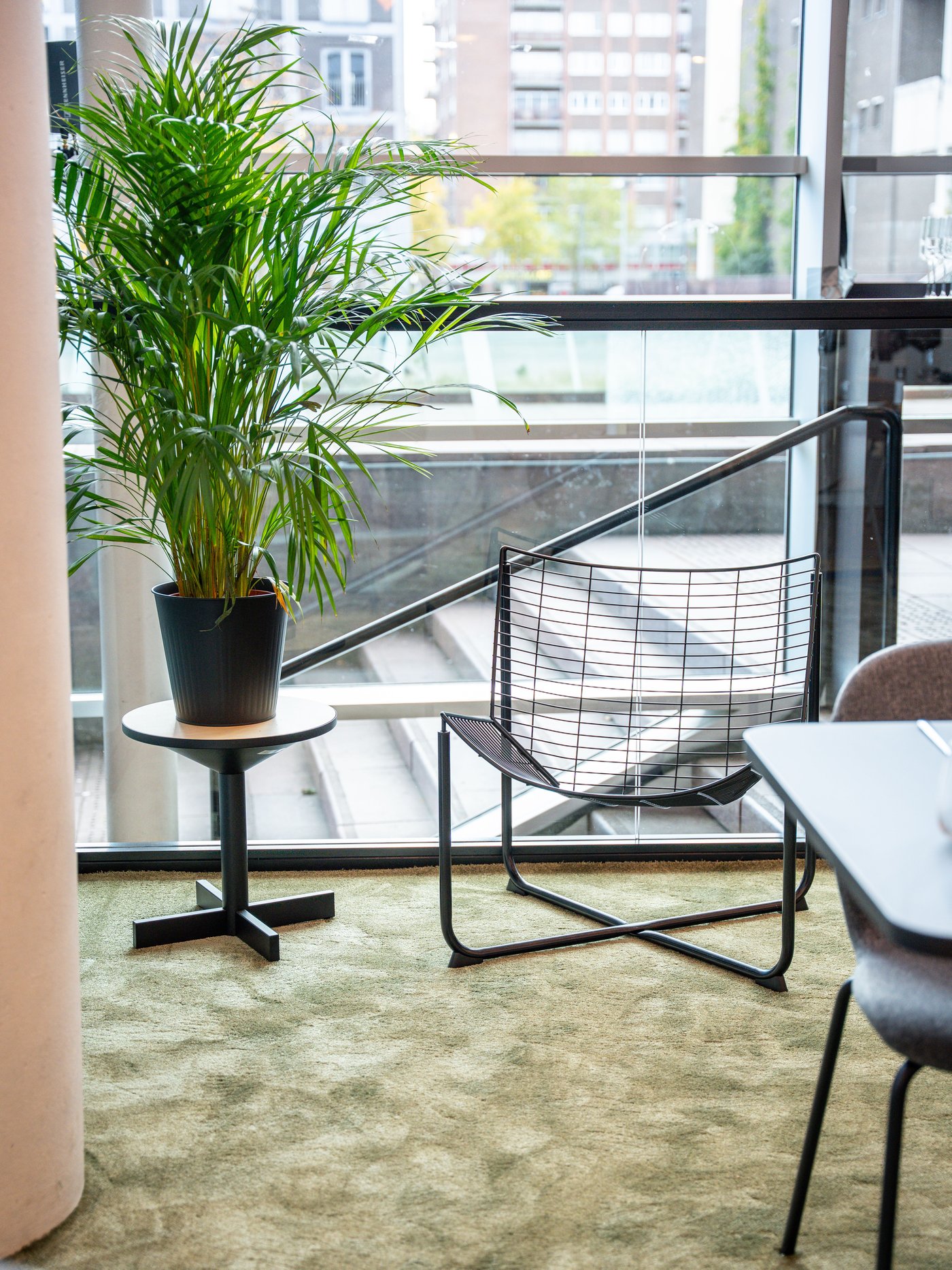 Zwarte draadstoel naast plant op tafeltje bij groot raam. Groen tapijt en zicht op stad zorgen voor moderne, luchtige sfeer in lounge of kantoorruimte.