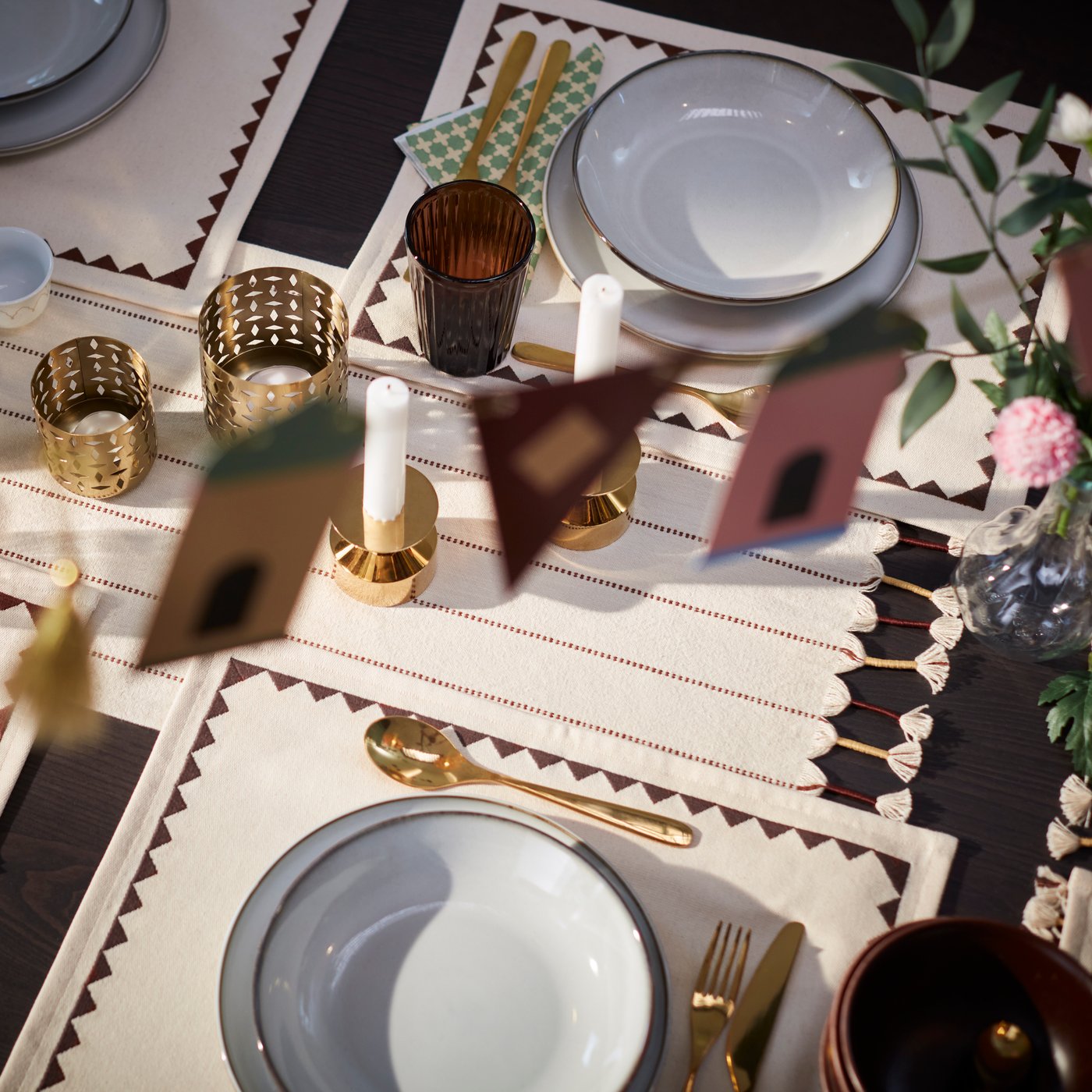 A beautifully set dining table, with an ornamented textile GOKVÄLLÅ tablerunner, gold-coloured candle lanterns and tableware.