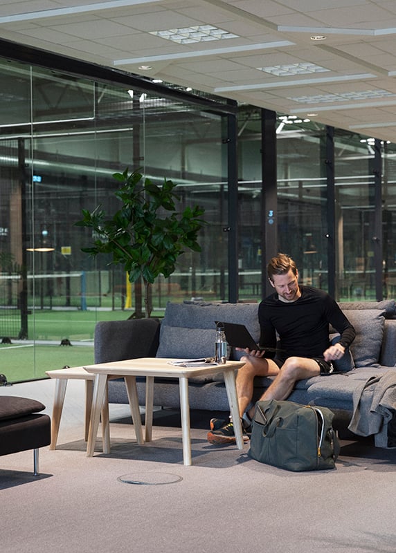 Person som sitter i en grå SÖDERHAMN soffa med en laptop på Birsta padelcenter. I bakgrunden skymtas padelbanor. 