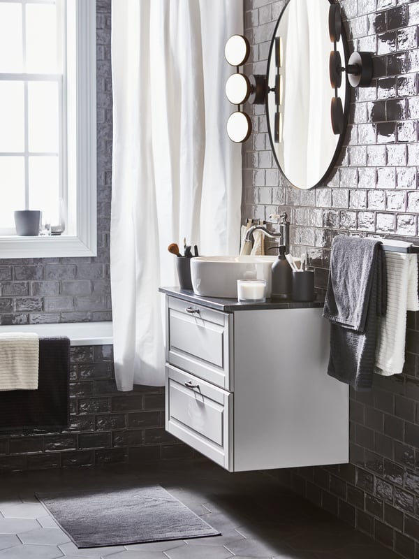 Une salle de bain avec des serviettes et draps de bain blancs et gris foncé, un rideau de douche BJÄRSEN blanc et un miroir rond noir.