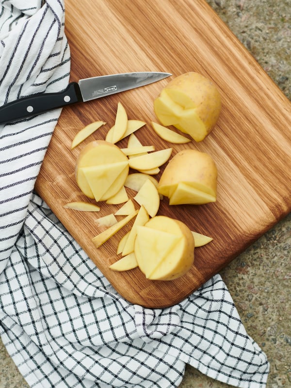 Chopped potatoes and a dark grey VARDAGEN paring knife atop an oak ARTISTISK chopping board, next to a RINNIG tea towel.