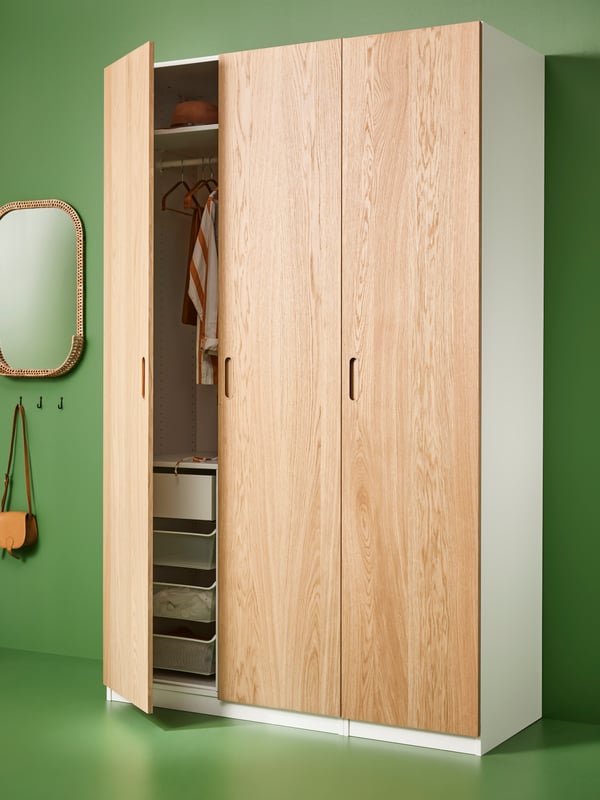 Blick auf einen PAX/TONSTAD Kleiderschrank mit Eichenfurniertüren, der neben einem SOMMARBO Spiegel an einer grünen Wand steht. Eine Tür des Schranks steht offen.