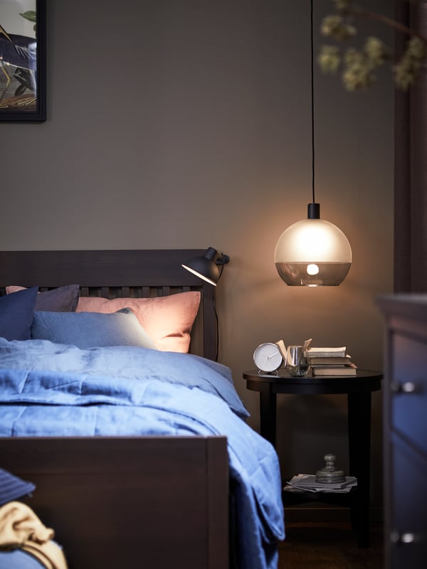 An IDANÄS bedframe with a PUDERVIVA dark blue quilt cover and pillowcases, next to a side table with a pendant light above.