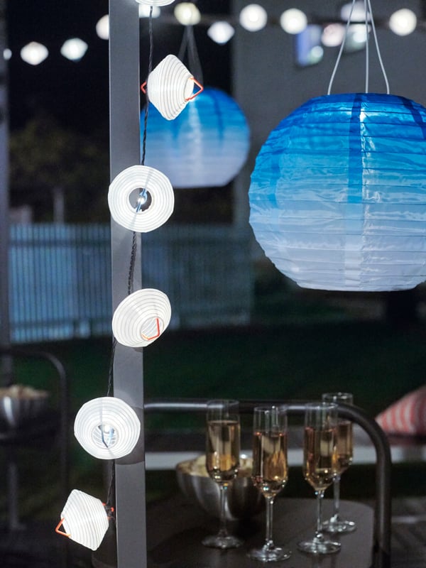 Blue-toned solar-powered SOLVINDEN LED pendant lamps, white lighting chains, wine glasses and snacks in a garden at night.