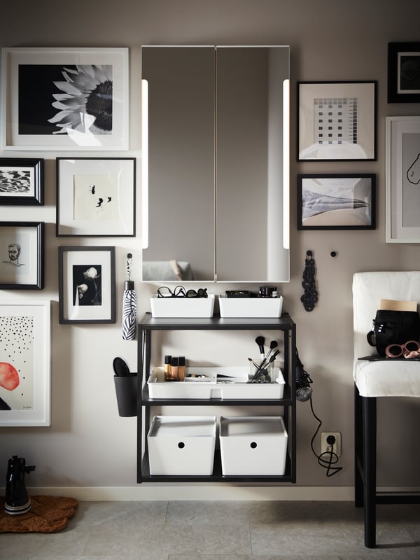 A white STORJORM mirror cabinet with built-in lighting on a wall with framed pictures and an anthracite shelving unit.