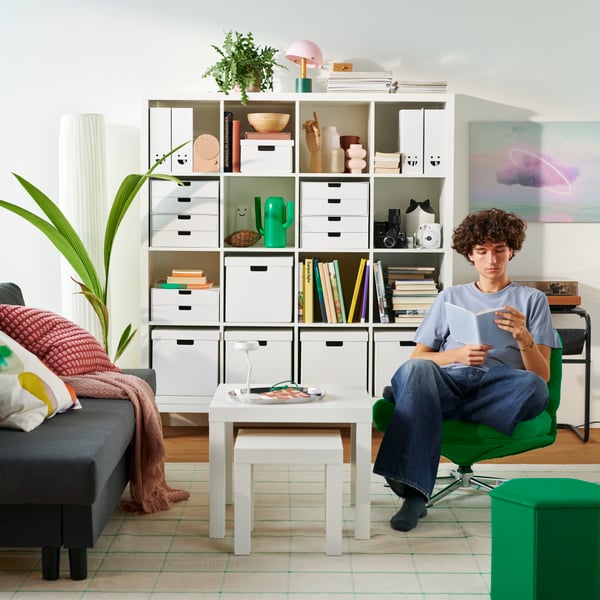 Ein Wohnzimmer mit einem KALLAX Regal, gefüllt mit Büchern und Dekoartikeln. Eine Person sitzt lesend in einem Sessel.