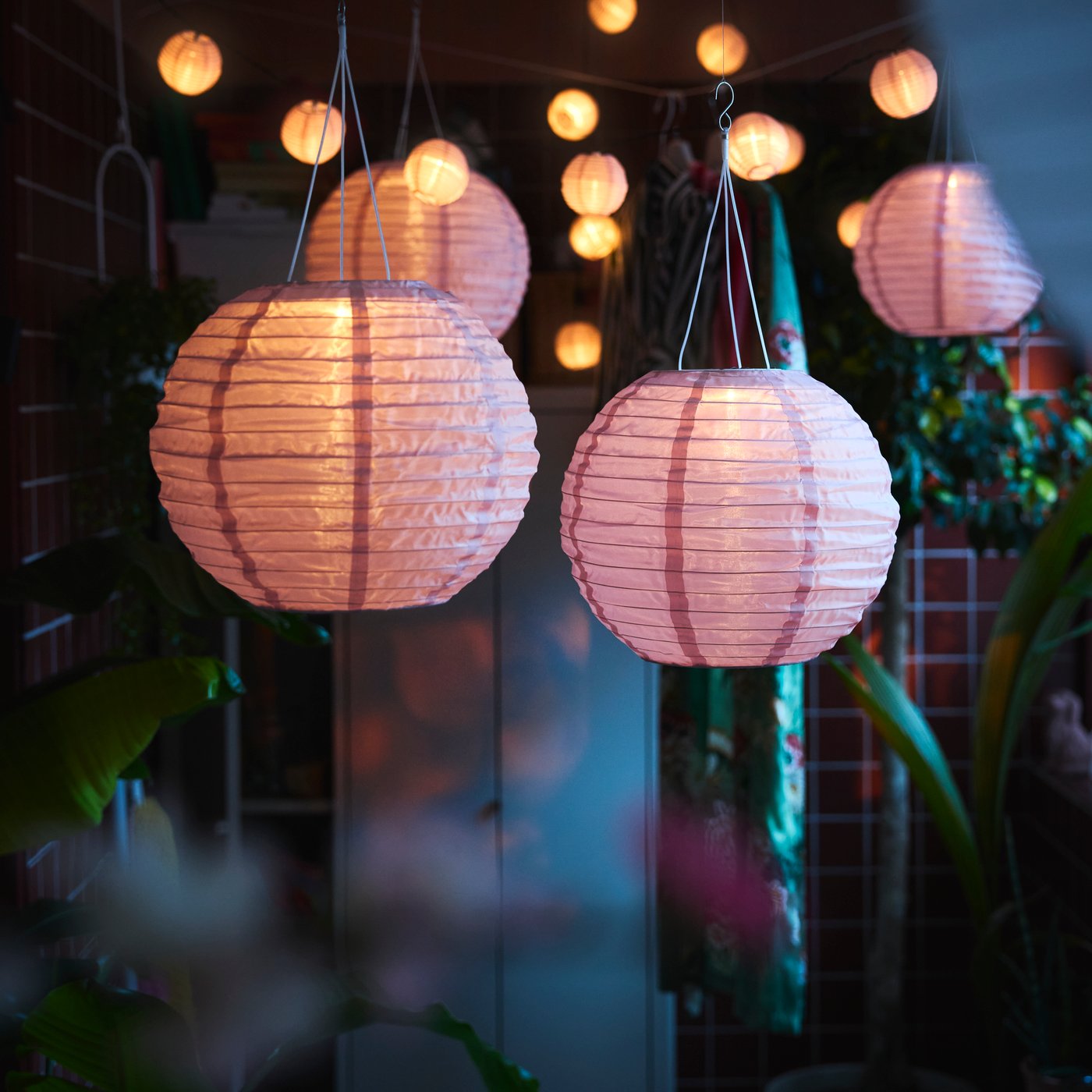 A balcony decorated with lush plants features several pink SOLVINDEN LED solar-powered pendant lamps hanging above.