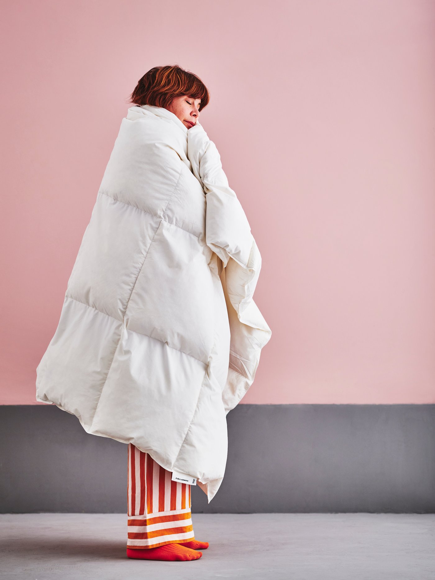 Person wrapped in a white down comforter stands against a pink wall, wearing striped pajama pants and orange socks.
