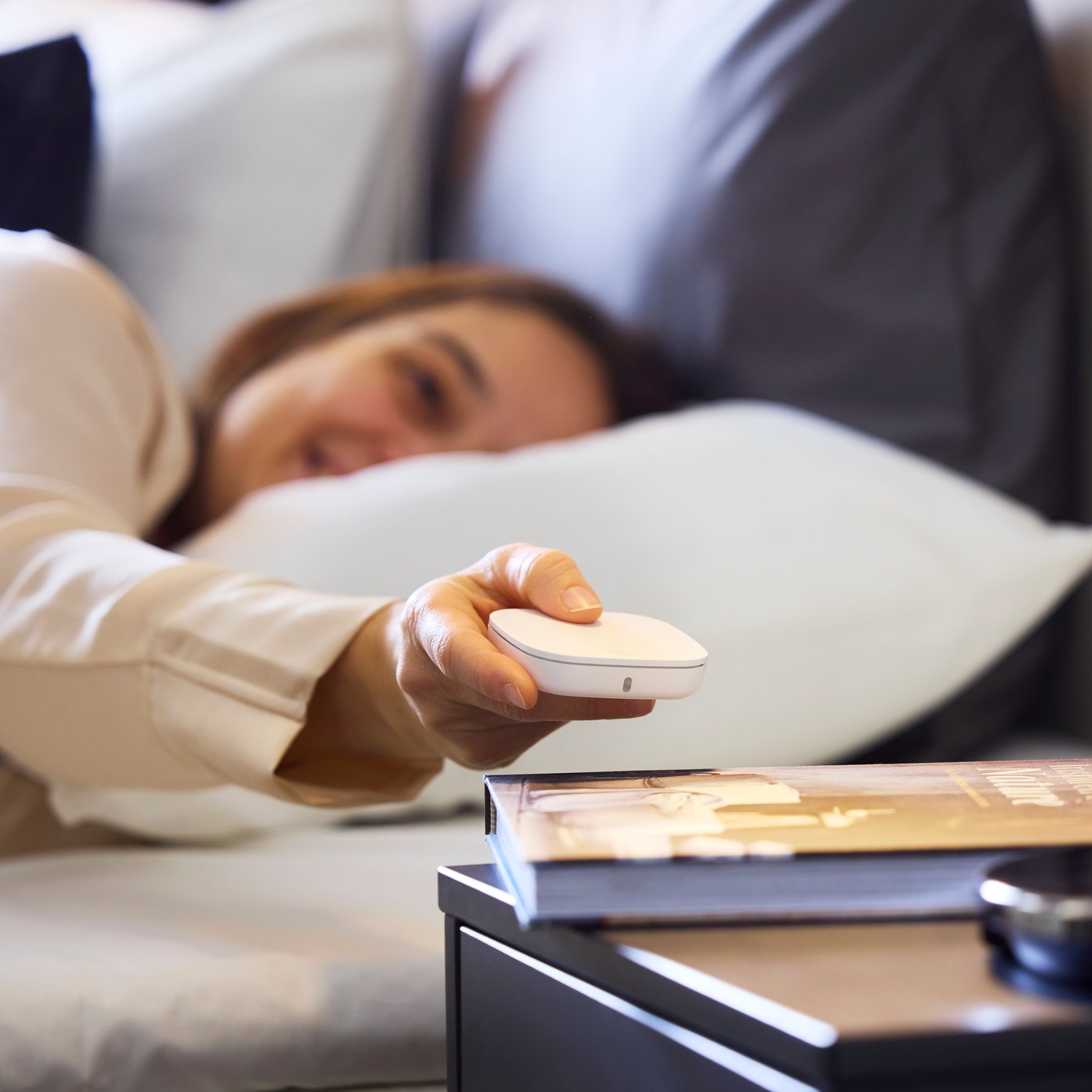 A person in a hotel bed holds a white STYRBAR remote, that is square with rounded corners, to control IKEA smart lighting.