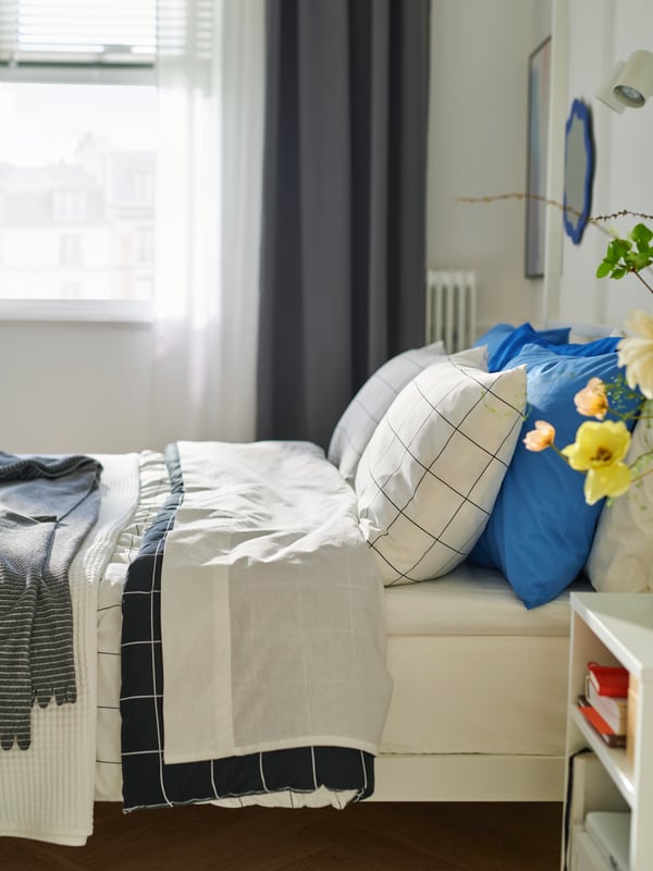 View of bed from the side with white bedding and blue pillows. Window in the background, with dark grey curtain at the side.