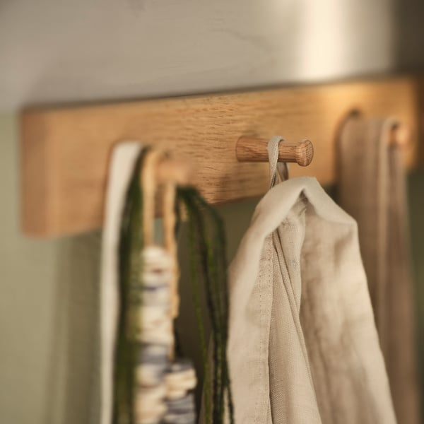Eine HÖVOLM Leiste mit 6 Knöpfen an einer Wand, u. a. mit Ketten und einer beigefarbenen Jacke.