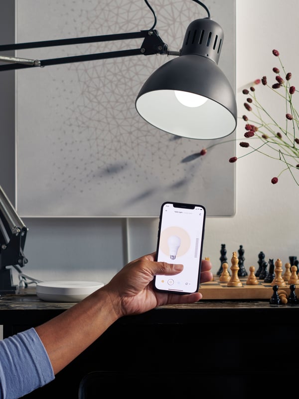 Osoba kontroliše lampu preko aplikacije IKEA Home smart na pametnom telefonu. Na stolu iza se vidi DIRIGERA baza.