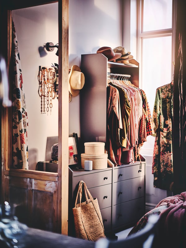 A grey HAUGA open wardrobe with three drawers, containing clothes and hats, set next to a grey chest with drawers.