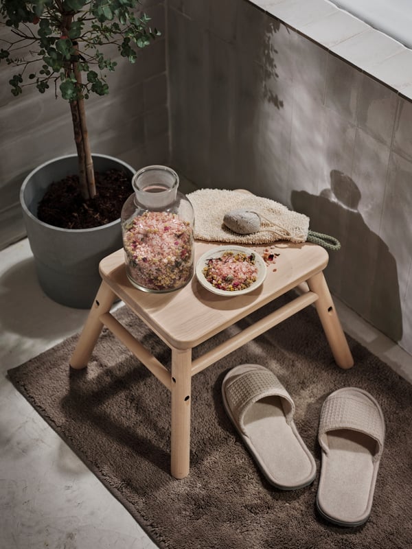 A scrub mit and scrub mix sitting on top of a wooden VILTO stool, with grey slippers next to it