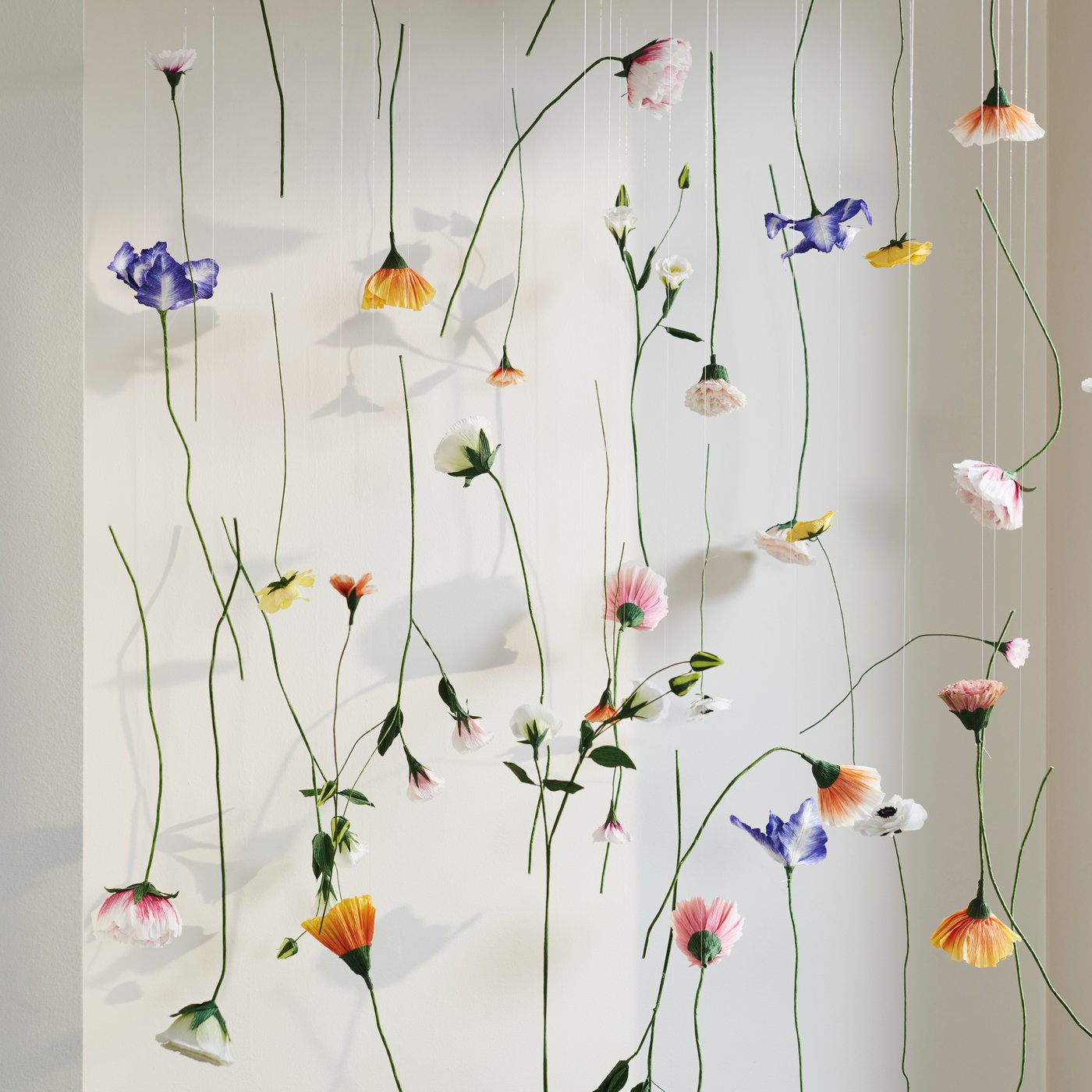 A lot of GULDSPETT artificial flowers are hung on different pieces of string to form a decoration in front of a wall.