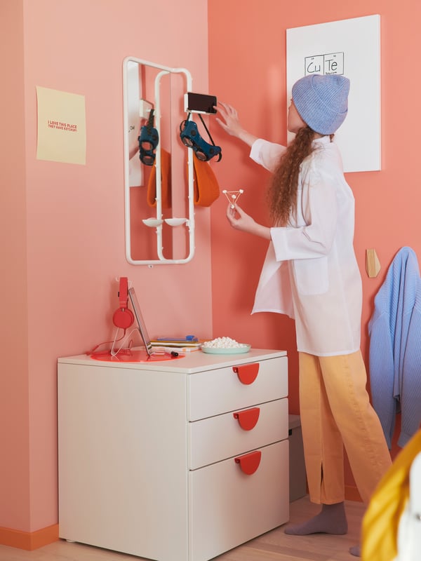 A girl standing by a white three-drawer SMÅSTAD/PLATSA chest adjusts a phone in the phone holder of a white MÖJLIGHET mirror.