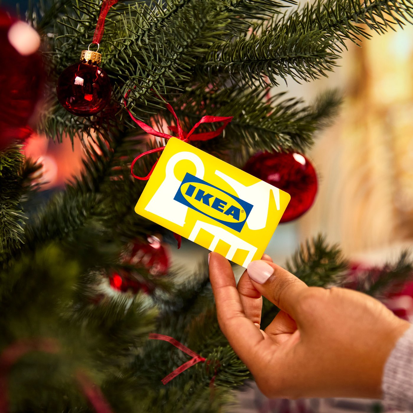 Eine Hand hält eine gelbe IKEA-Geschenkkarte vor einem geschmückten Weihnachtsbaum mit roten Kugeln und Schleifen. Die Szene vermittelt eine festliche, fröhliche Atmosphäre.