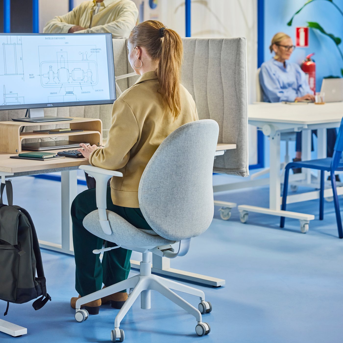 Person seated at a desk using a computer with technical drawings, partition panels and coworkers at laptops in a blue office.