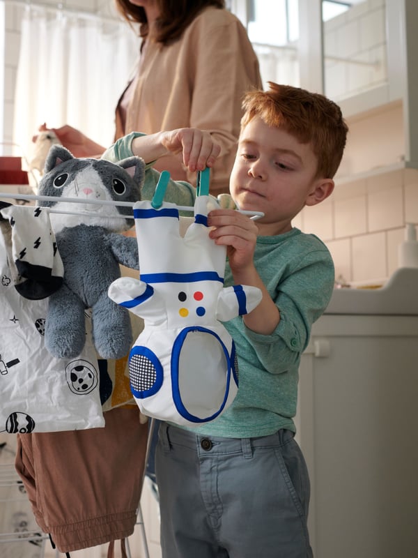 Standing next to their parent, a child hangs a small spacesuit onto an indoor washing line. A soft toy cat is hangs there.