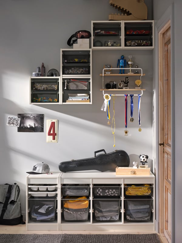 A number of white TROFAST frames on the floor and wall filled with grey storage trays and mesh storage boxes holding clothes.