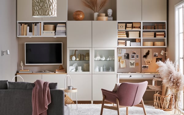 Salon moderne avec rangement sur mesure, étagères ouvertes et espace de travail intégré. La pièce comprend une télévision, des objets décoratifs, un fauteuil rose, un canapé gris et une palette de couleurs neutres et chaleureuses.